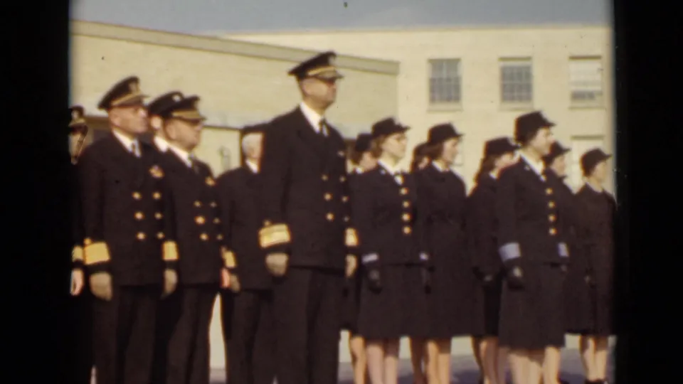 A tall uniformed officer stands among sailors and servicewomen in a formal outdoor formation, with windows visible behind them.
