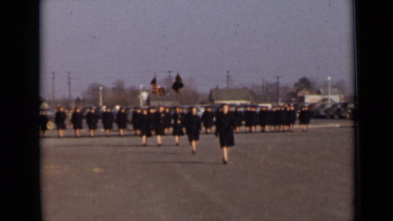 Women's Army Corps members appear to march in formation on a paved New York lot, led by flag bearers carrying the American flag and a dark standard.
