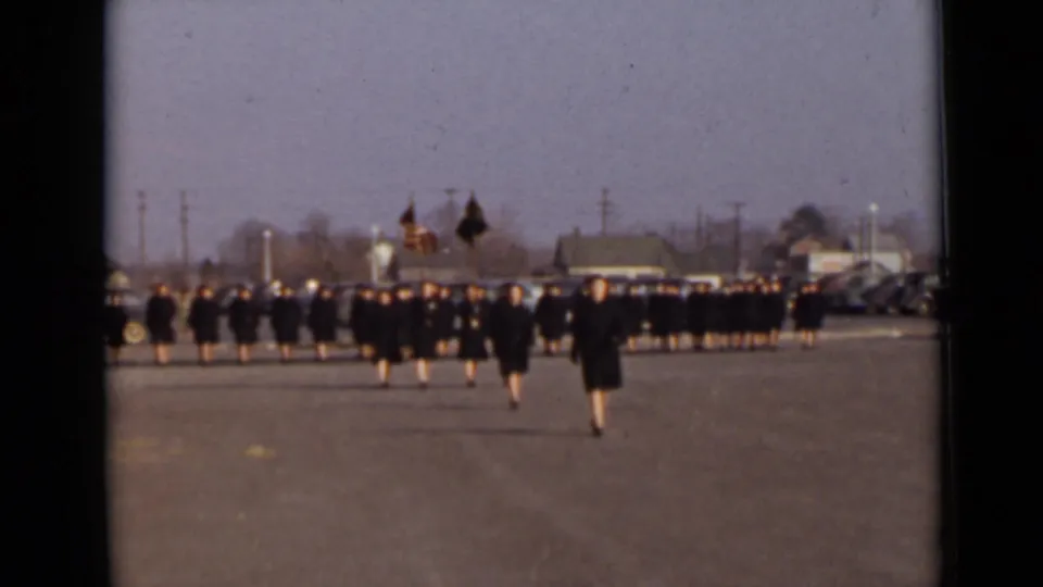 Women's Army Corps members appear to march in formation on a paved New York lot, led by flag bearers carrying the American flag and a dark standard.