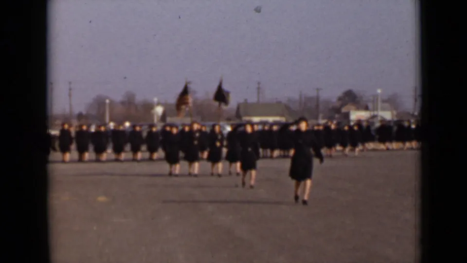 Women in dark military uniforms move in ordered rows across the paved area, with flags and a larger formation visible behind them.