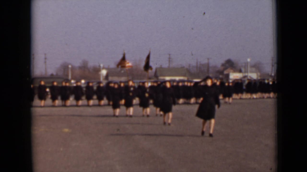 Women in dark coats march in rows across a paved field with flags behind them.
