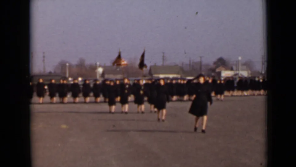 In 1947 New York, women in matching dark coats march in formation across a paved field while flag bearers stand or move behind them.