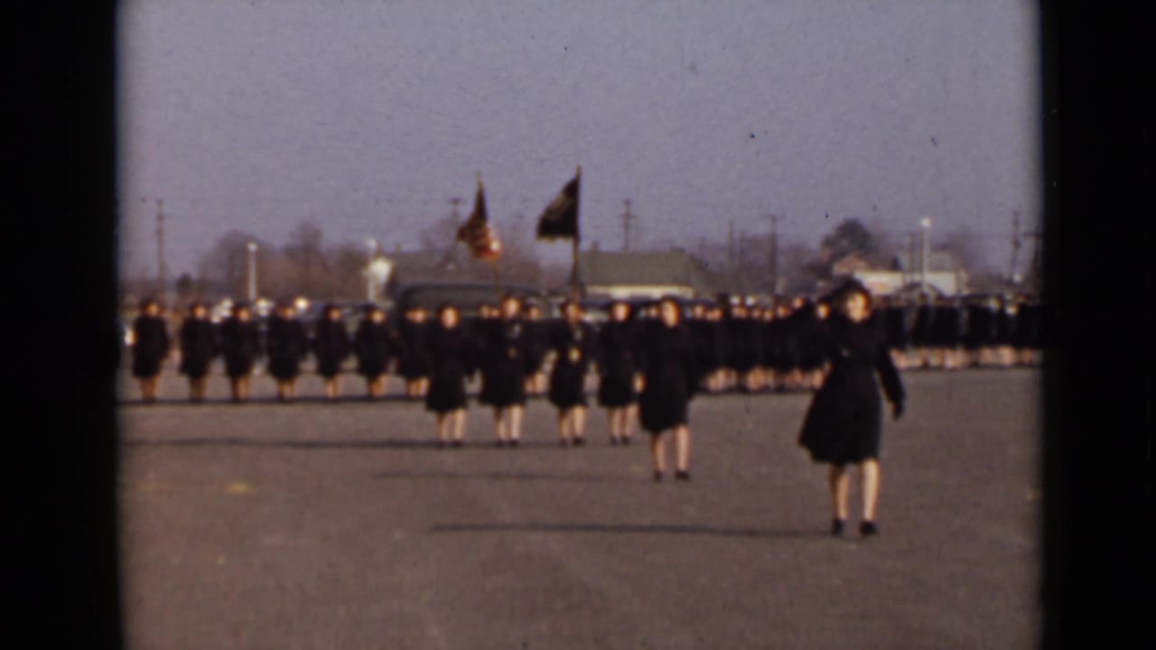 A formation of women in dark coats marches toward the camera on a wide paved area.