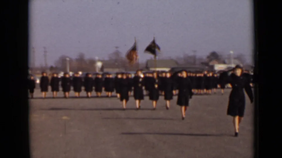 A wide 8mm frame shows women in dark coats marching in line in New York, with flag bearers and distant buildings behind the formation.