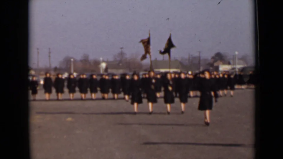 The closing frame shows rows of women in dark coats marching on a paved New York field, with two flags and a larger formation behind them.