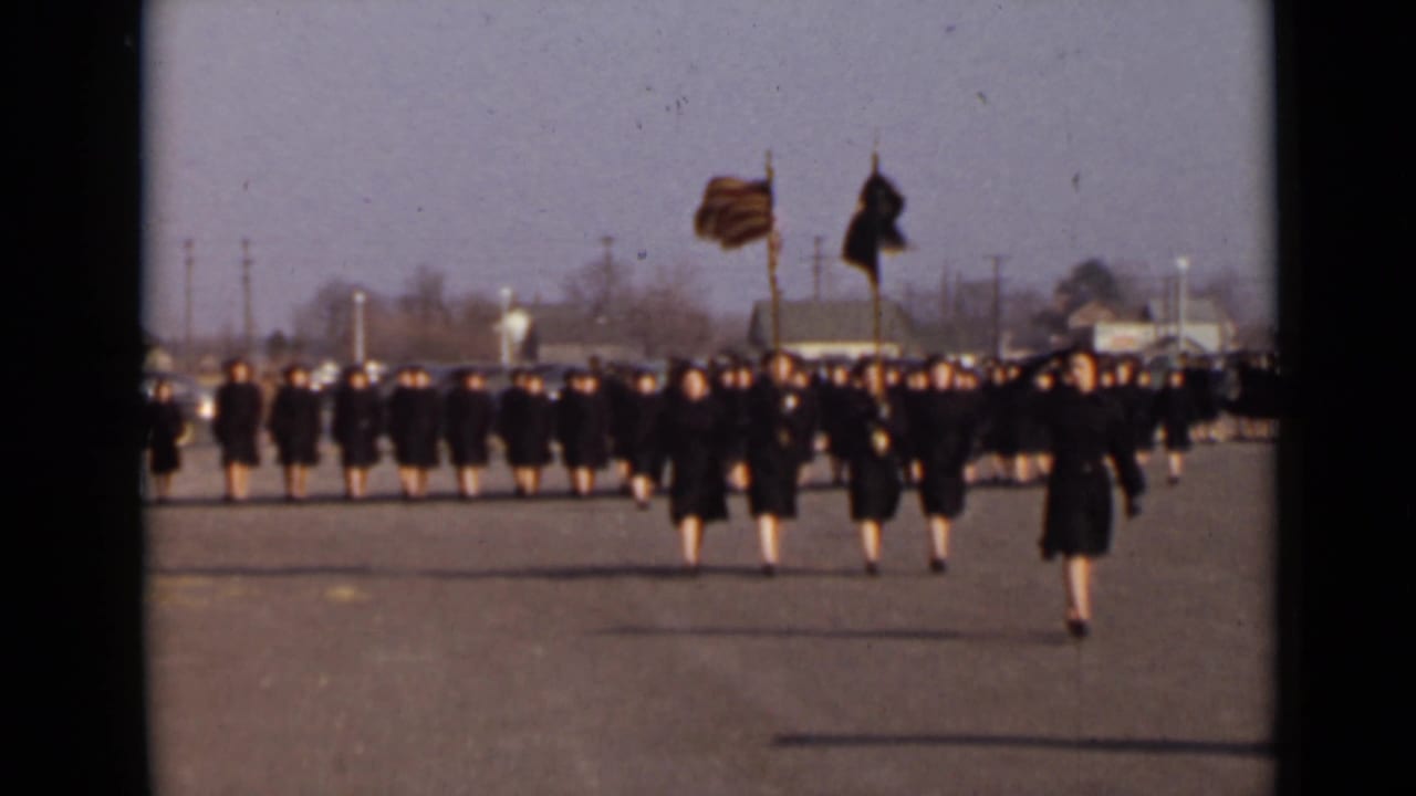 Women in dark uniforms march in formation across a paved lot with flags visible ahead.