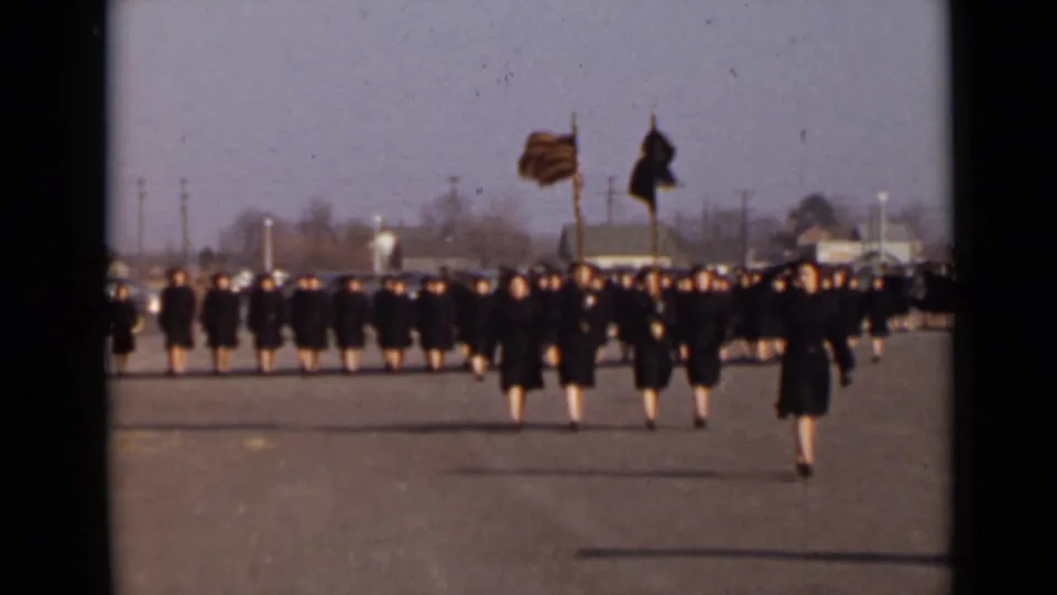 1947 New York home-movie frame showing women in dark uniforms marching toward the camera in a formal formation with flags in front.
