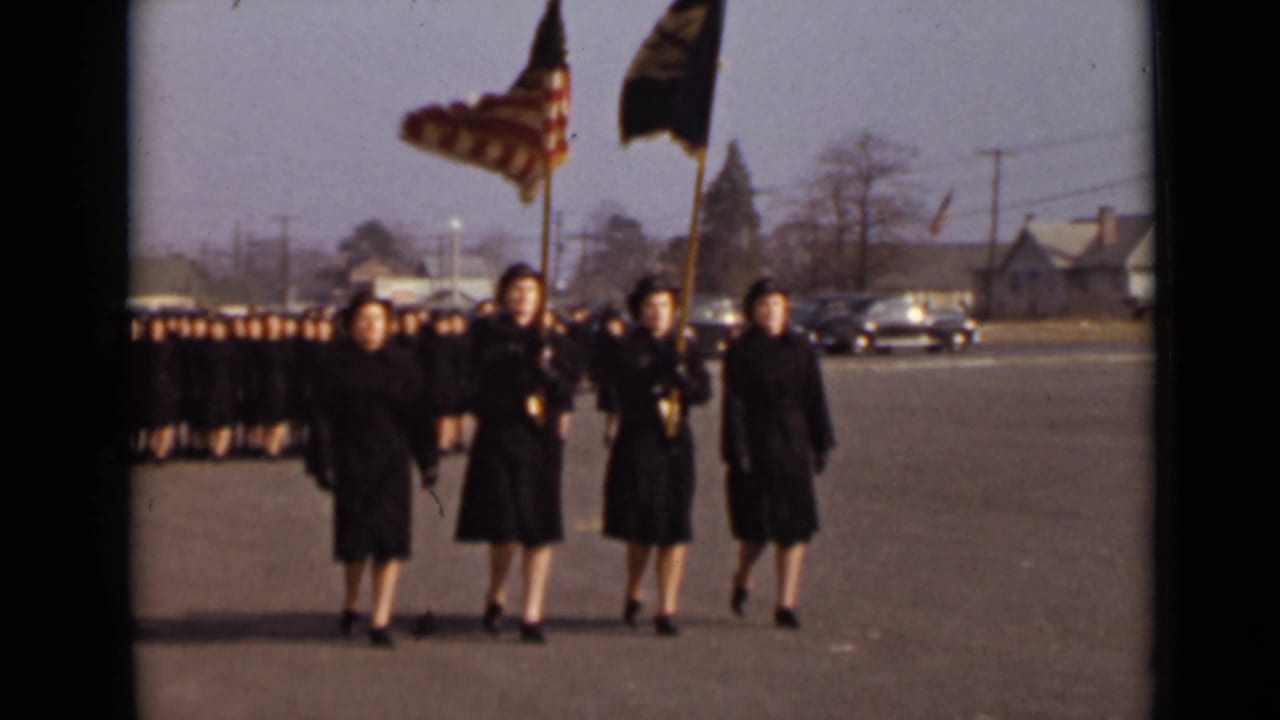 Four women in dark uniforms advance at the front of a larger marching group with two flags.