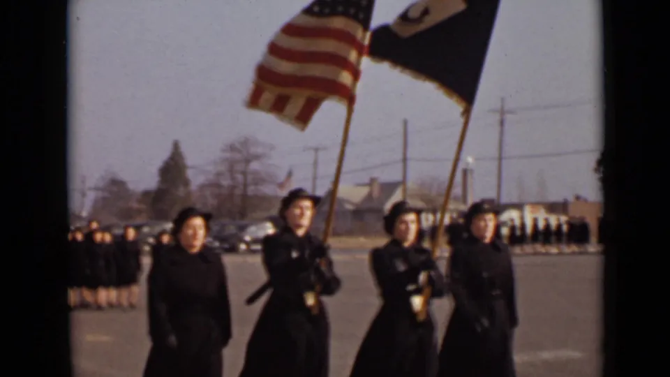 1947 New York home-movie frame of women in dark uniforms marching under an American flag and dark banner, with formation rows behind them.