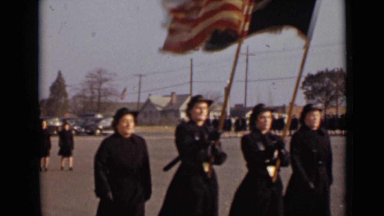 In 1947 New York footage, women in matching dark formal coats and hats lead a procession while two front figures carry flags.