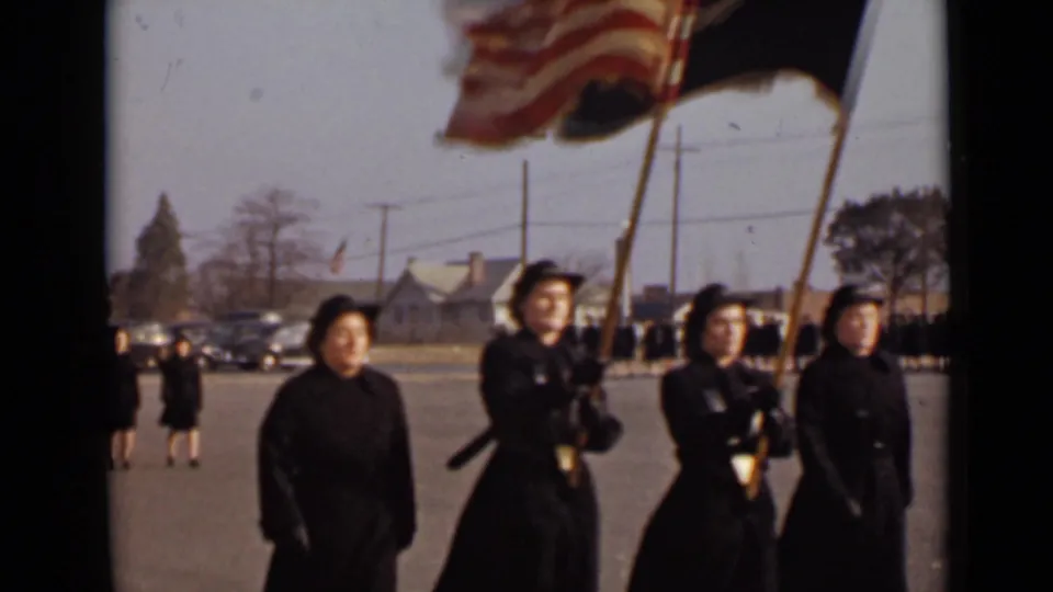 In 1947 New York footage, women in matching dark formal coats and hats lead a procession while two front figures carry flags.