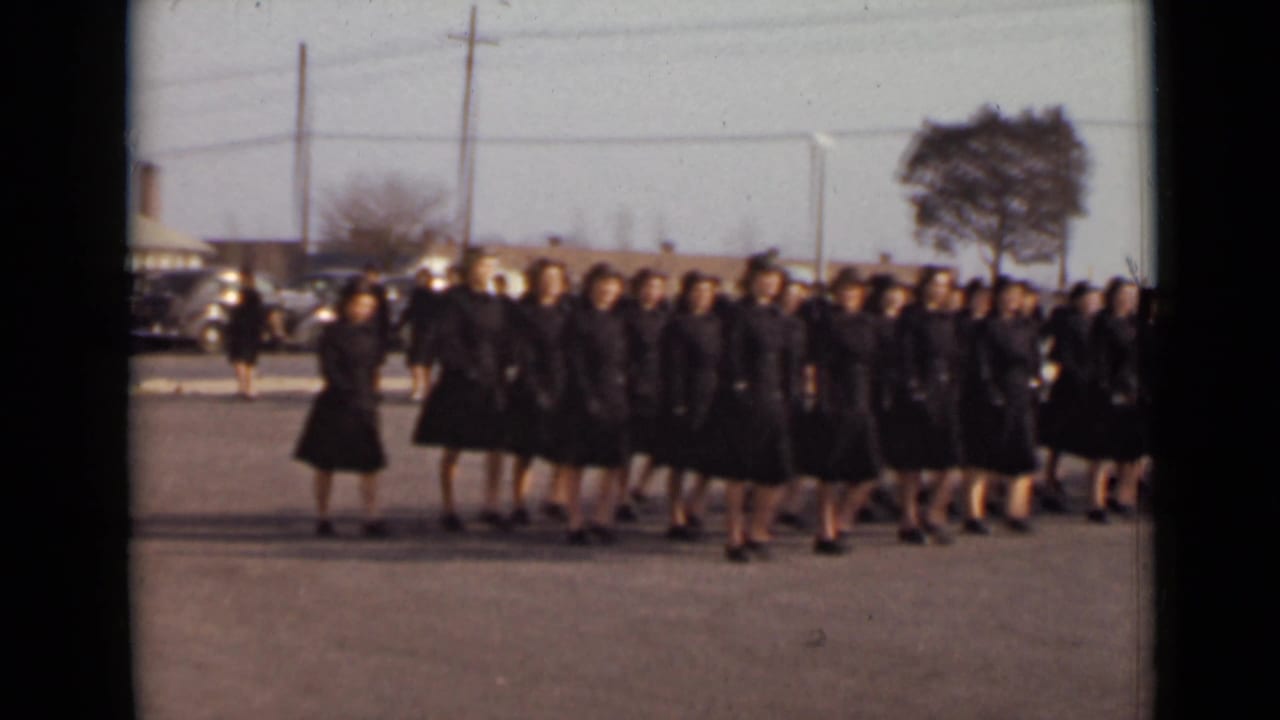A large women’s marching group moves in formation across a paved open area in archival 1947 New York home-movie footage.