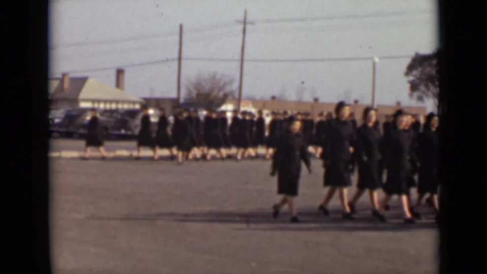 Women in matching dark coats pass through the foreground while additional participants remain lined in the background of the 1947 New York scene.