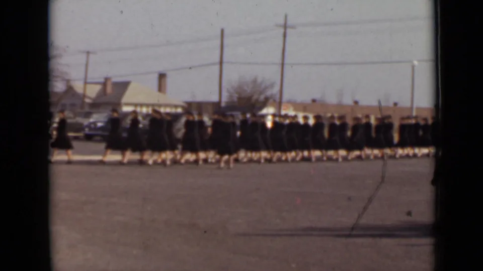 A long formation of women in dark formal clothing crosses the open paved area, with cars and low buildings in the background.