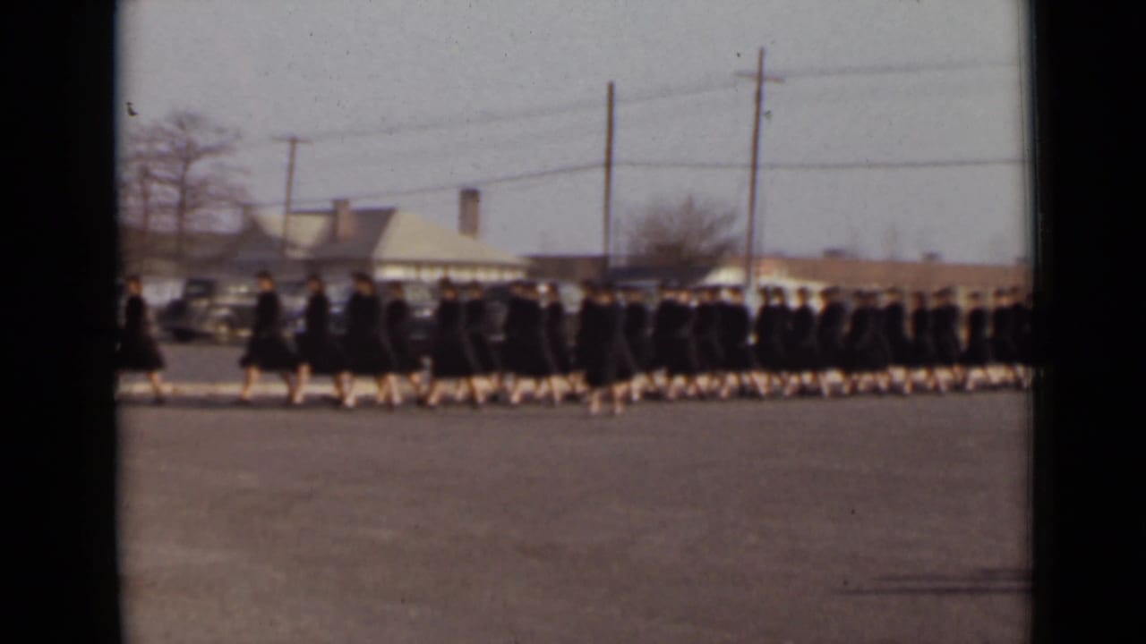 Graduates in dark gowns march across a paved area with parked cars and utility poles behind them.