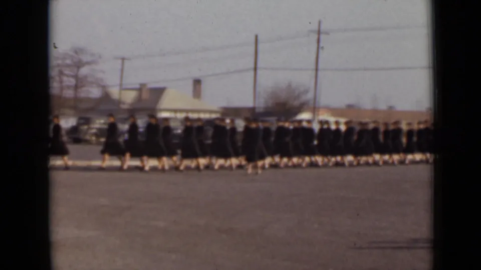 1947 New York home-movie frame showing young women in black graduation gowns and caps walking in formation past parked cars and low buildings.