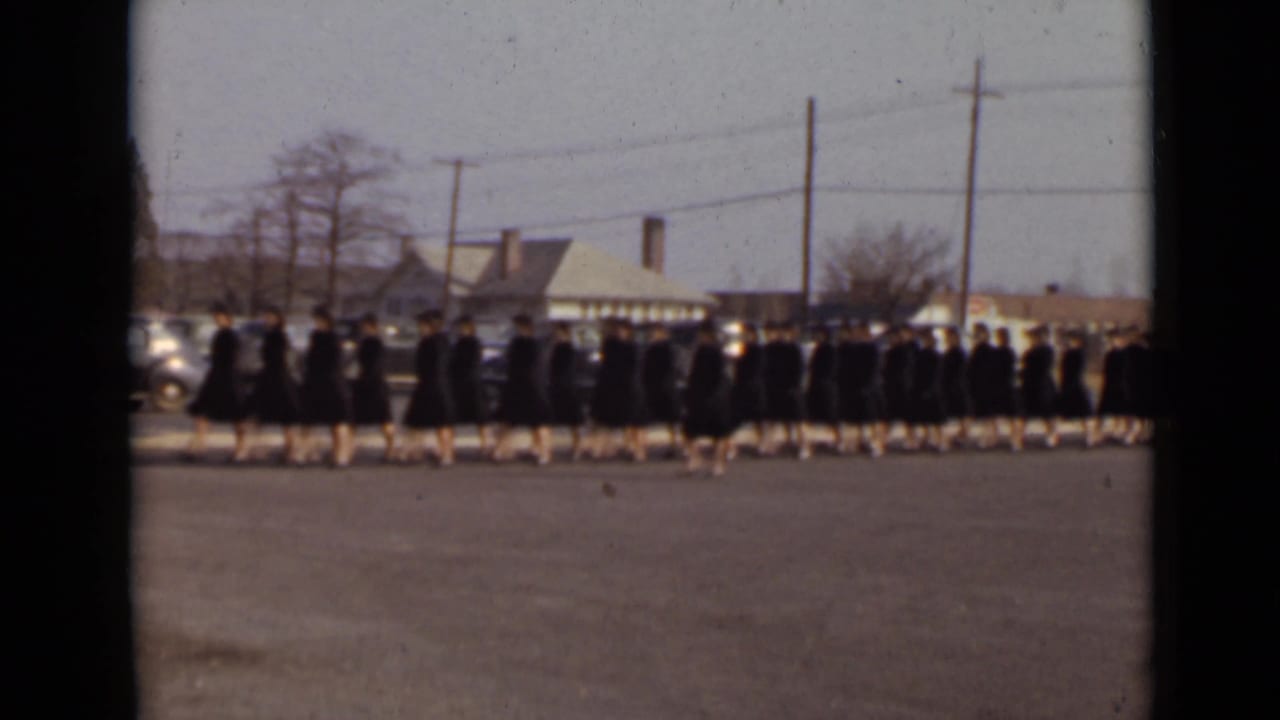 Line of graduates in dark gowns continues past houses, cars, and utility poles.