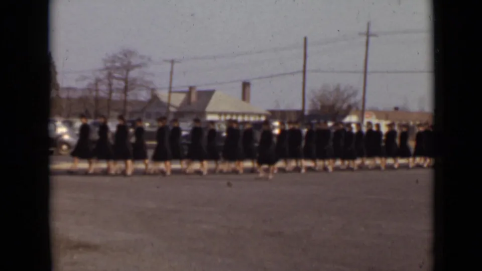 1947 New York graduation procession with students in black gowns and caps moving across an open paved field against a suburban backdrop.