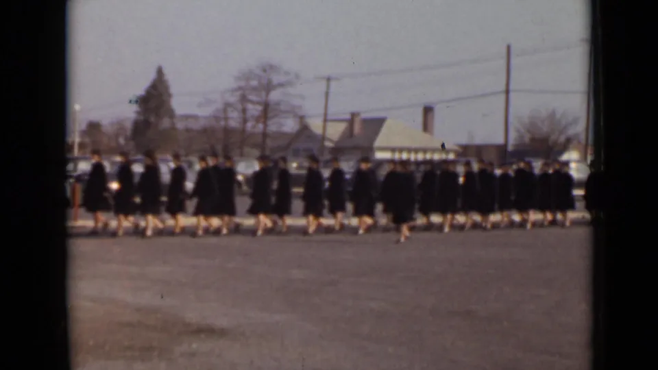 1947 New York home-movie view of a graduation line moving across pavement, with dark gowns, caps, parked cars, and modest buildings visible.