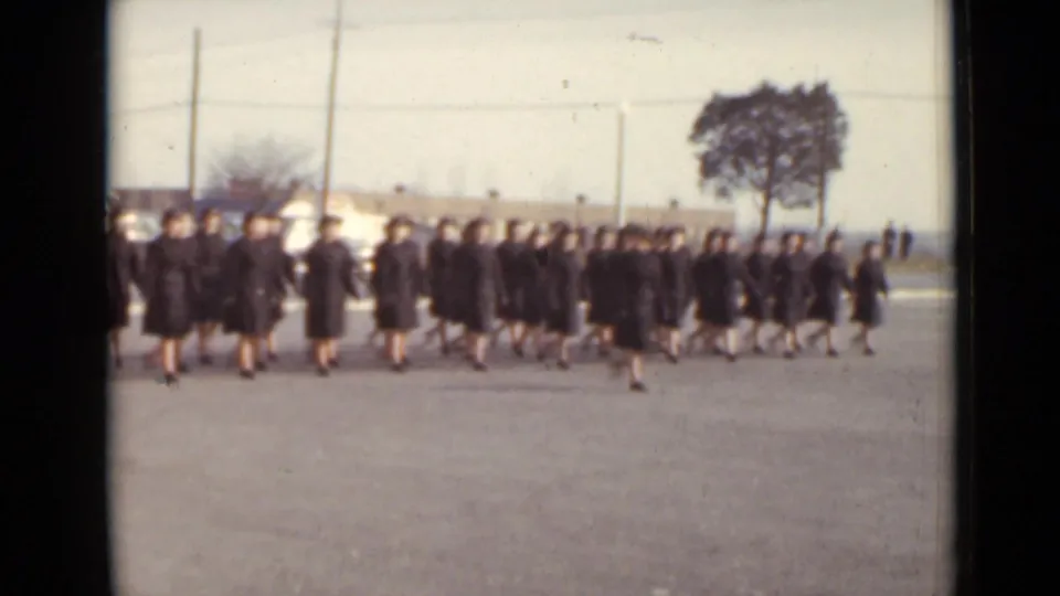 1947 New York graduation scene showing a cluster of students in black gowns and caps crossing bright pavement with cars, poles, and a tree behind them.