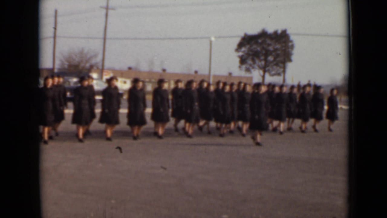 Women in matching dark outfits march across a paved lot in formation.
