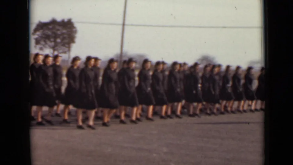 1947 New York home-movie frame of women in dark coordinated dress marching left to right across an open paved lot.
