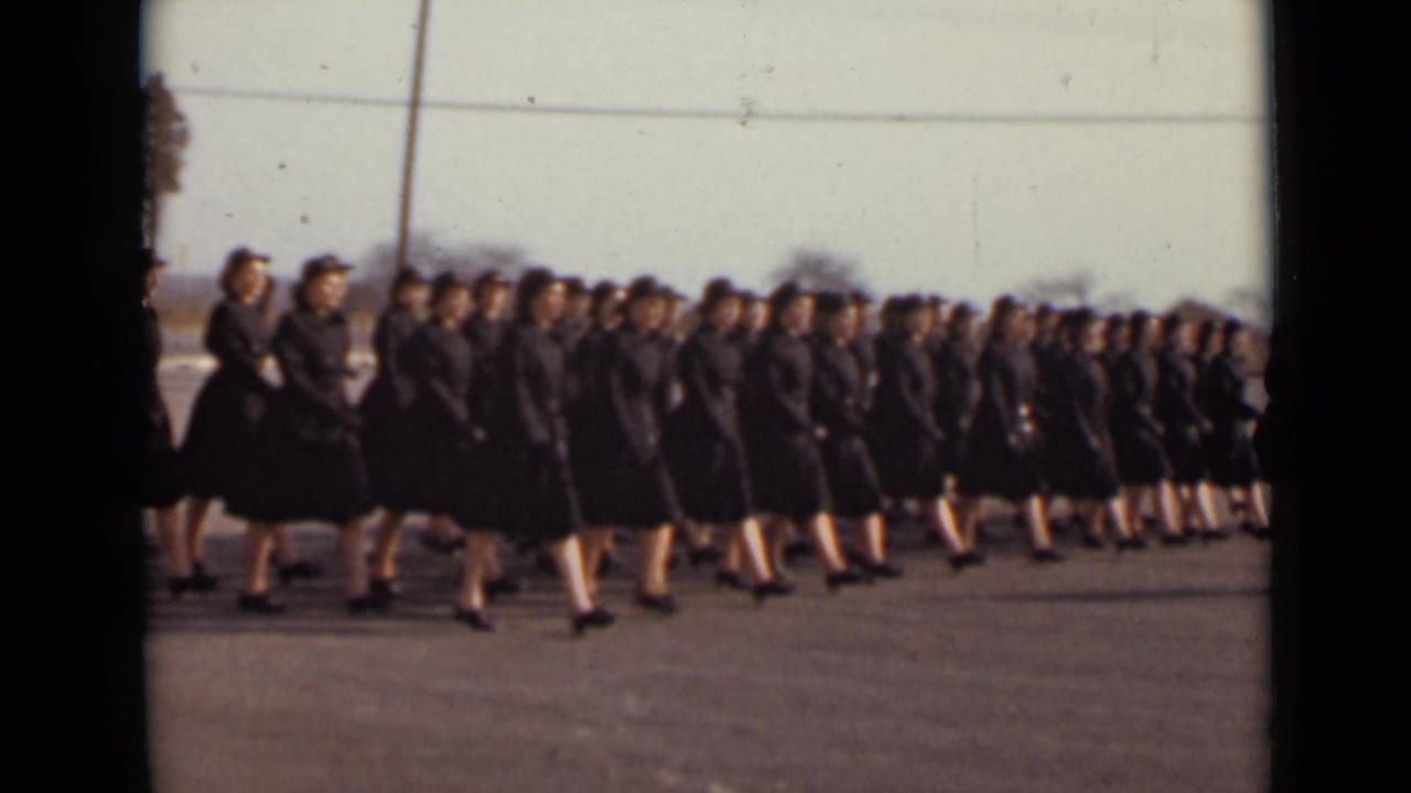 Rows of women in dark uniforms march across an open paved area.