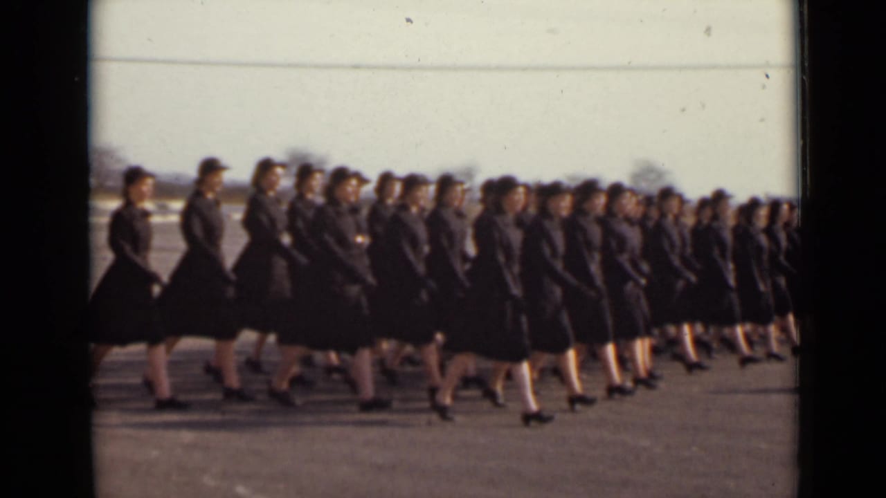 A large group of women in dark mid-century uniforms and hats moves in step across a paved New York setting.