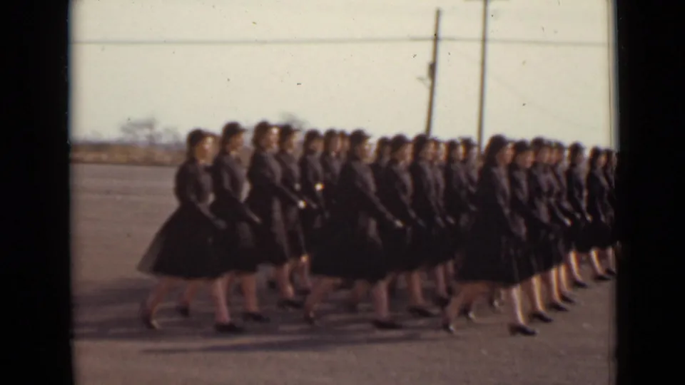 The handheld 8mm view follows rows of women in dark uniforms marching across open pavement in 1947 New York.
