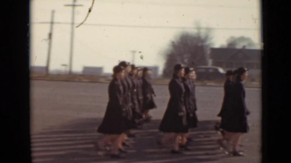 Uniformed women march in smaller groups at the edge of the formation, with low buildings and utility poles in the background.