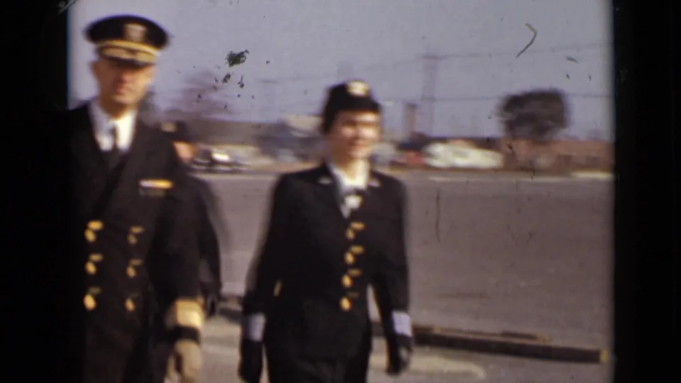 A male officer and a uniformed woman walk across a paved area in 1947 New York home-movie footage.