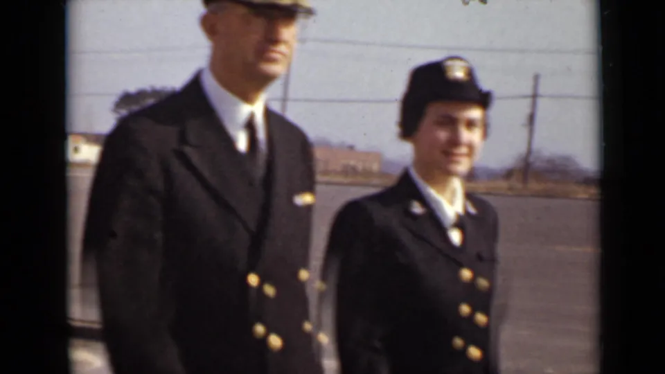 A male officer and a woman in dark uniform face the camera in a paved outdoor area, with distant buildings behind them.