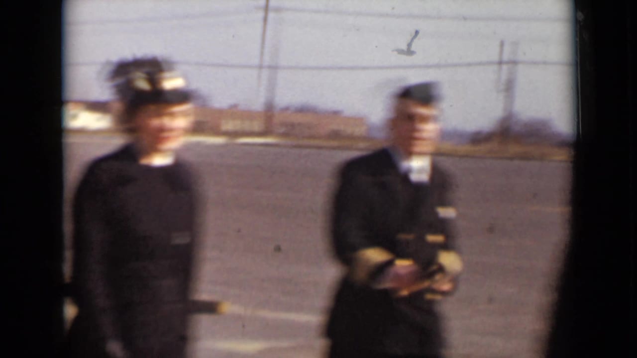 Two uniformed women standing on a paved open area with a brick building behind them.