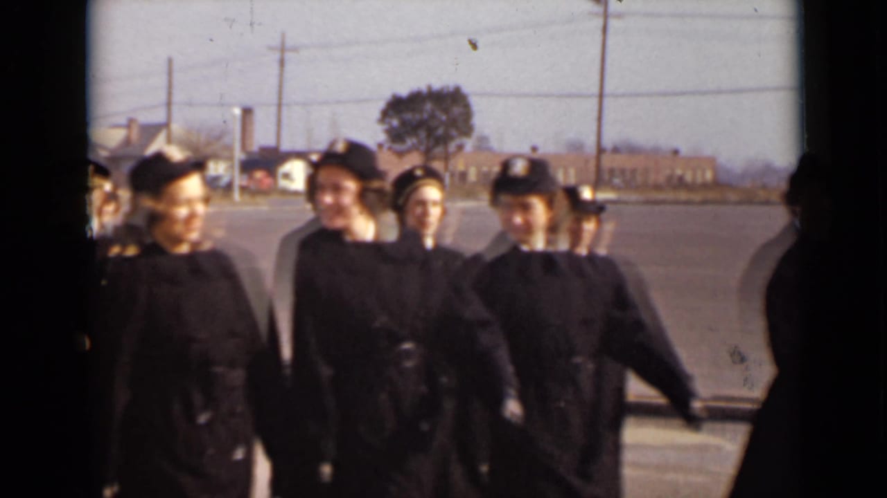 Uniformed women in dark caps and coats walk across a paved lot.