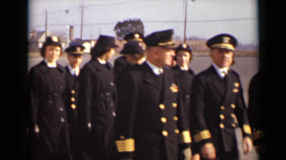 Uniformed women and naval-style officers continue past the camera in the paved-lot setting, with faces and insignia partly visible.