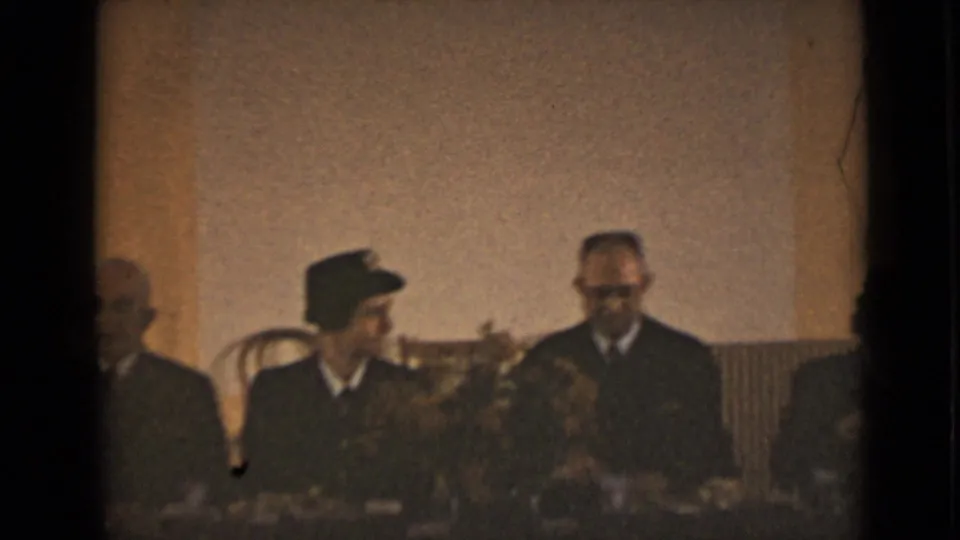The scene shifts indoors, where uniformed and formally dressed adults sit at a table with flowers visible in the foreground.