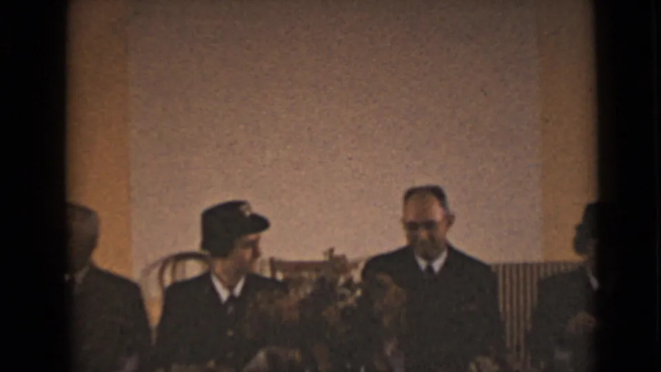 Uniformed and formally dressed adults remain seated at an indoor table, facing one another in a quiet social moment.
