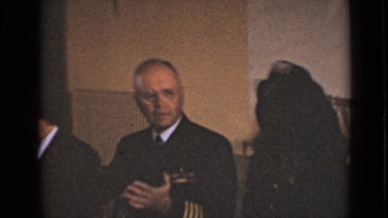 Older man in dark uniform with epaulets stands indoors near other guests.
