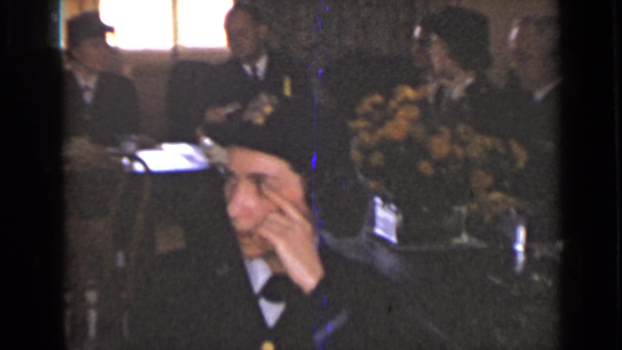 Woman in a dark cap adjusts her face or hat while seated near flowers and guests.