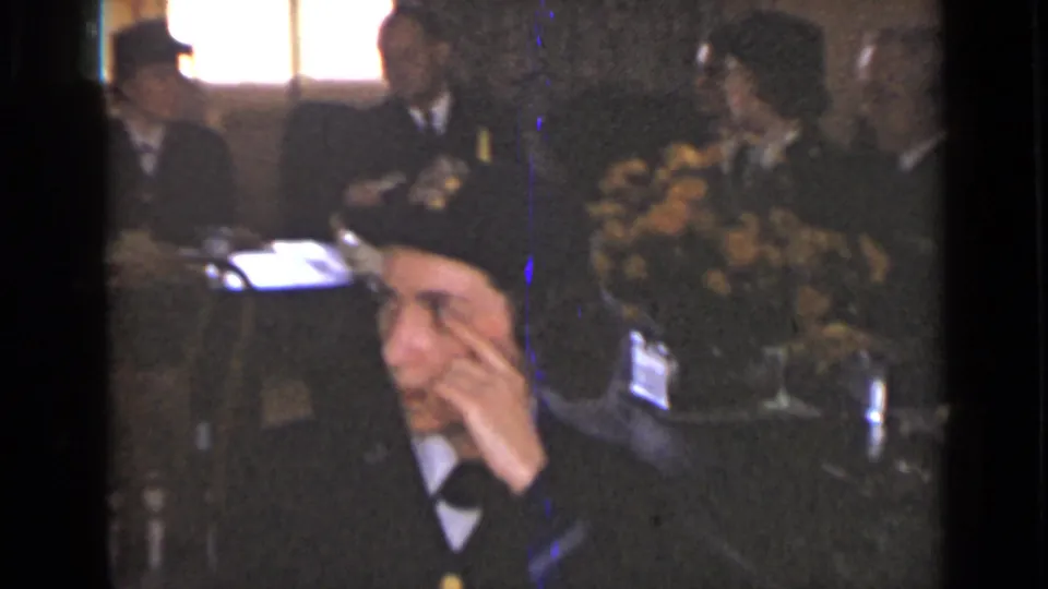 A woman in a dark uniform-style cap sits at a dinner table in the 1947 New York footage, with floral arrangements and other guests behind her.