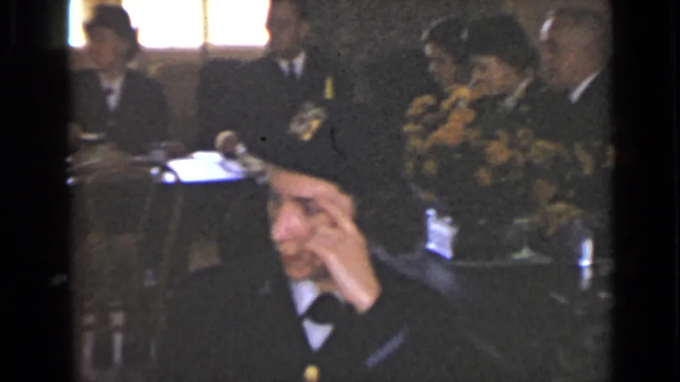 The 8mm frame shows a woman in a dark cap in the foreground, with men in formal clothing, flowers, and table items in the private dinner setting.