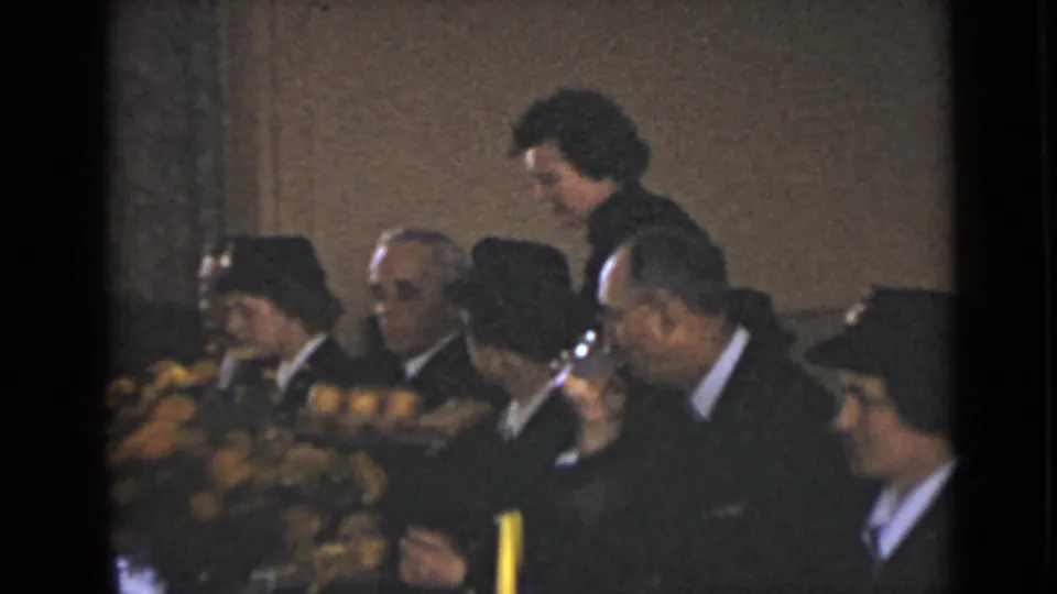 A wider 1947 New York home-movie frame shows adults gathered around a long dinner table with flowers, a candle, and several dark uniforms or formal outfits.