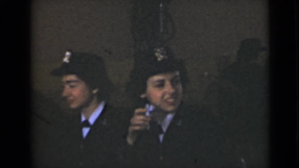 Two women in dark military-style uniforms sit in a dim New York interior, one raising a small object near her face.