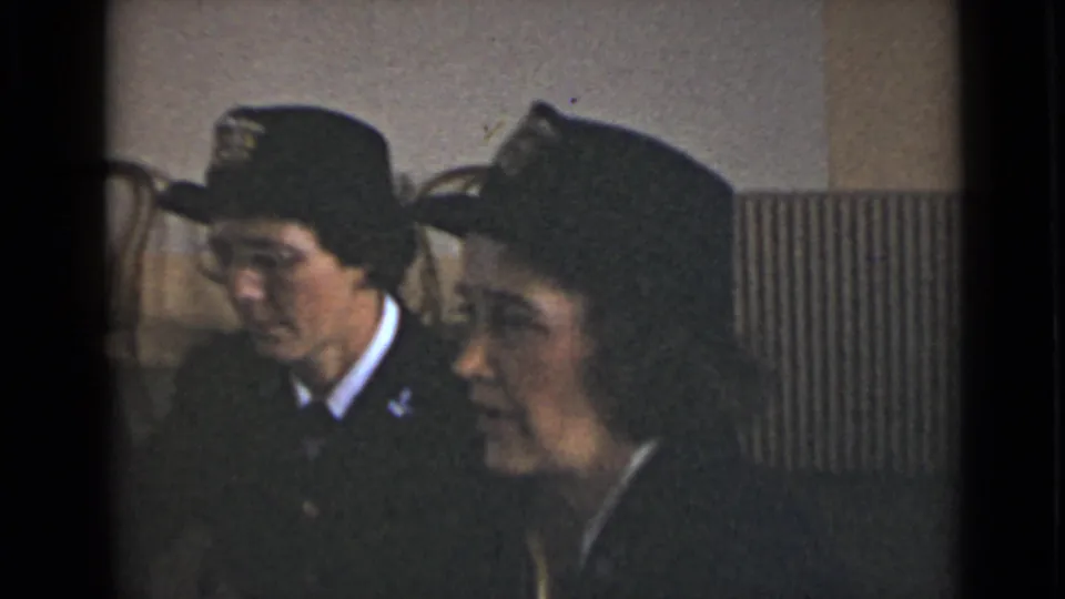A uniformed woman sits in profile beside another person in a brighter interior with chairs and a radiator behind them.