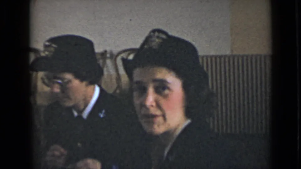 A woman in a dark uniform and cap faces the camera in a bright interior, with another uniformed person seated behind her.