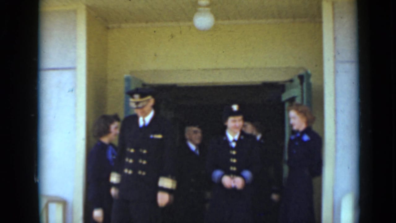 Uniformed men and women stand at a building entrance with open doors behind them.
