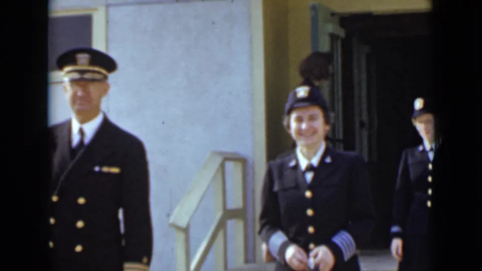 A woman in a dark military-style uniform walks toward the camera as other uniformed personnel emerge from the doorway behind her.