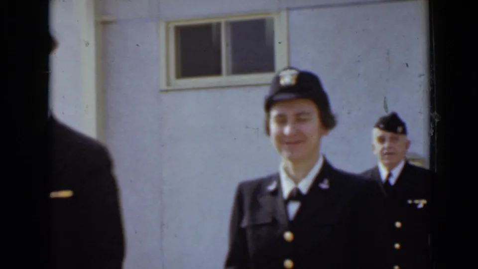 A woman in a dark uniform and cap is framed near the building wall, with another uniformed person visible in the background.