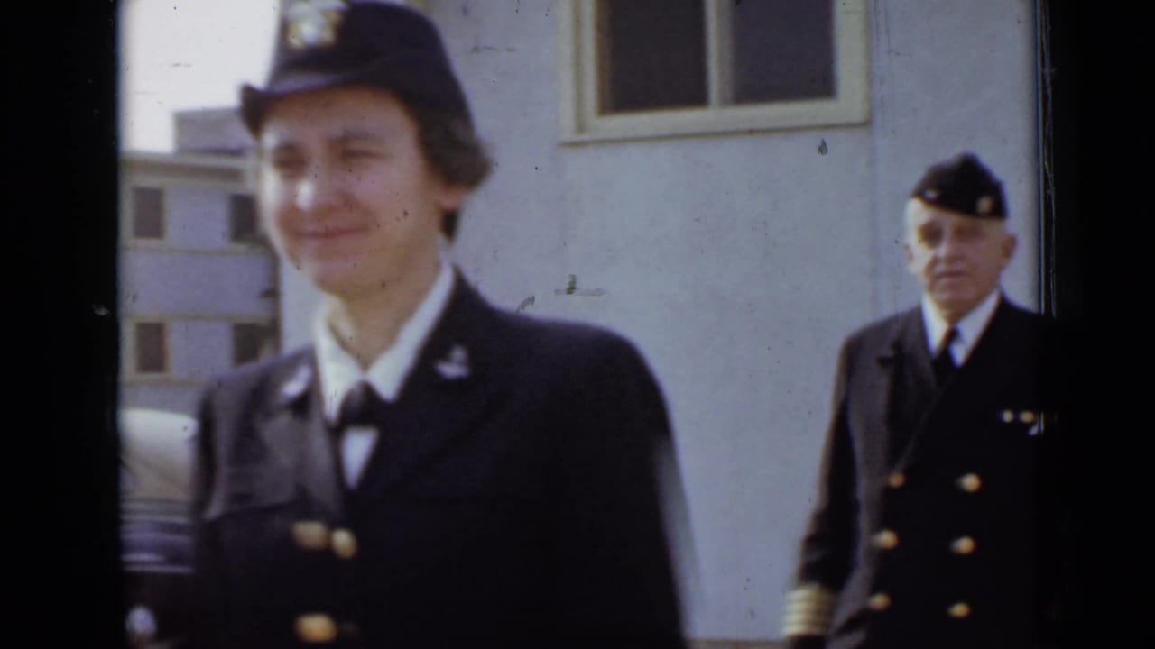 Woman in a dark uniform and cap walks past a light-colored building with an officer behind her.