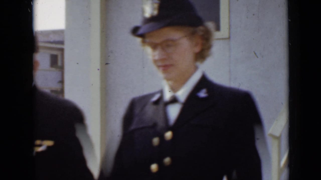 Woman in a dark uniform and cap walks beside a building, with a partial figure in the foreground.